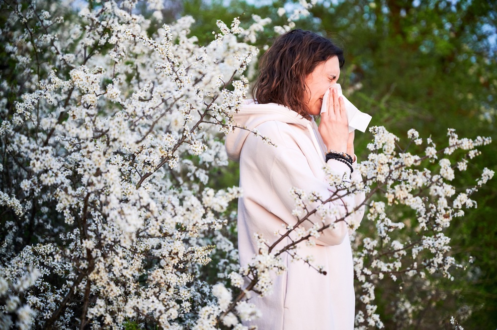 a woman under a flowering tree blowing her nose due to allergies
